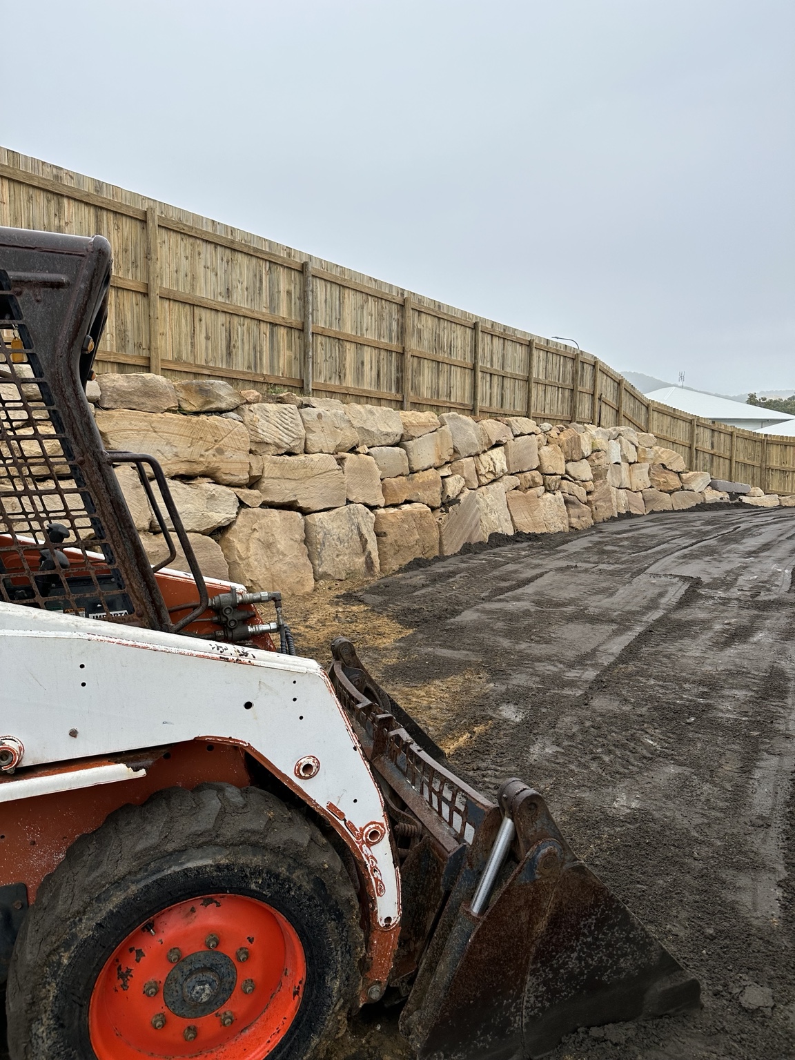 Bobcat spreading topsoil and preparing ground beside a sandstone retaining wall